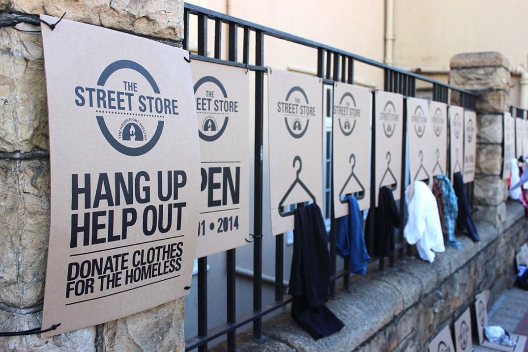 Street store cardboard hangers hanging on a rail, some with clothes attached to them. Text on the hangers reads 'Hang Up. Help Out. Donate Clothes for the Homeless'.